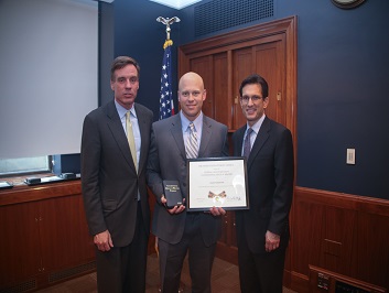 A group of three people standing together. The man in the middle is holding a Badge of Bravery award.