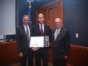 A group of three people standing together. The man in the middle is holding a Badge of Bravery award.