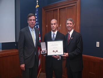 A group of three people standing together. The man in the middle is holding a Badge of Bravery award.