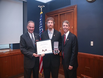 A group of three people standing together. The man in the middle is holding a Badge of Bravery award.