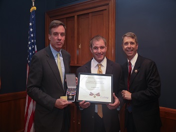 A group of three people standing together. The man in the middle is holding a Badge of Bravery award.