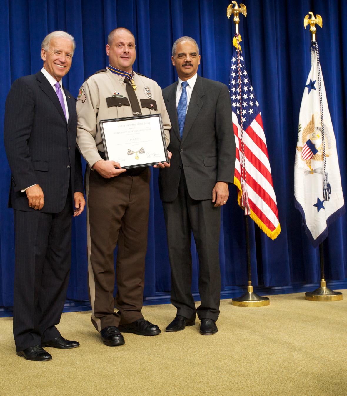 Three men standing in front of flags on a stage and facing the camera. The man in the middle is wearing an officer uniform and holding a Medal of Valor award plaque. 