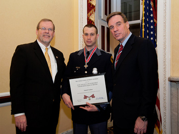 A group of three people standing together. The man in the middle is holding a Badge of Bravery award.