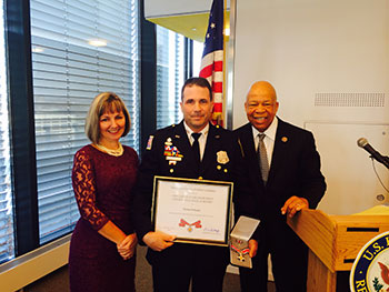 A group of three people standing together. The man in the middle is holding a Badge of Bravery award.