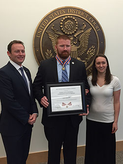 A group of three people standing together. The man in the middle is holding a Badge of Bravery award.