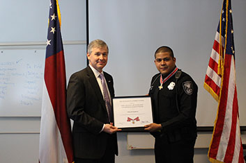 Two people standing together at the Badge of Bravery awards ceremony, holding an award plaque between them, and smiling at the camera