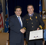 Two men standing on a stage, shaking hands, and smiling at the camera. One man is in an officer uniform and is holding a Medal of Valor award plaque.