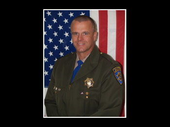 a portrait of a man in a sheriff uniform standing in front of an American flag and facing the camera