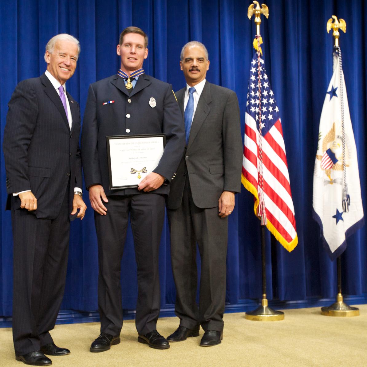 Three men standing in front of flags on a stage and smiling at the camera. The man in the middle is wearing an officer uniform and holding a Medal of Valor award plaque. 