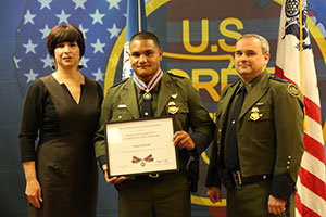 A group of three people standing together. The man in the middle is holding a Badge of Bravery award.