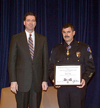 Two men standing on a stage and facing the camera. One man is wearing an officer uniform and holding a Medal of Valor award plaque.