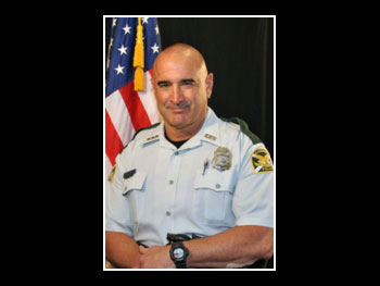 a head and shoulders portrait of a man in an officer's uniform facing the camera and standing in front of an American flag 
