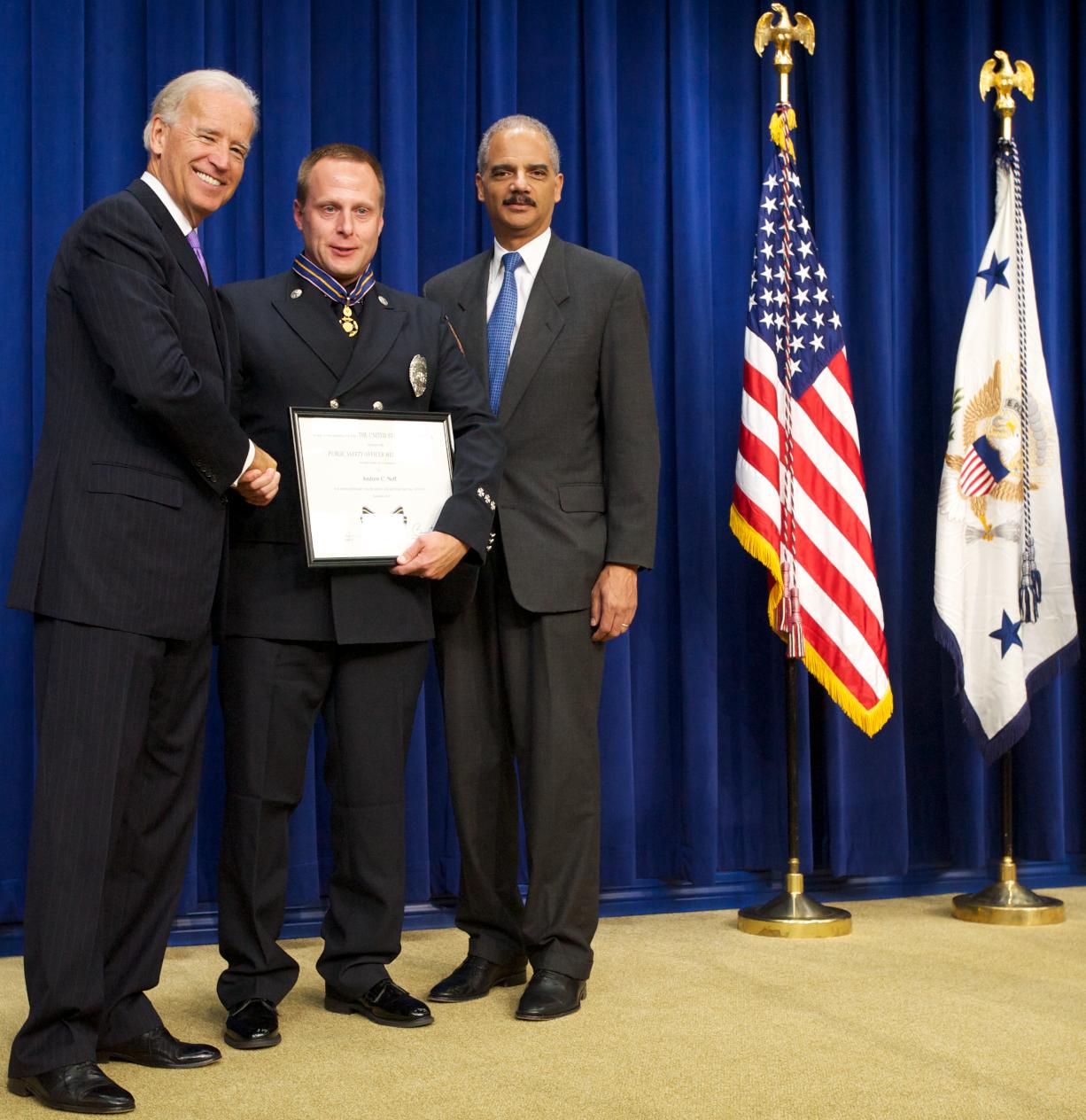 Three men standing in front of flags on a stage and smiling at the camera. The man in the middle is wearing an officer uniform and holding a Medal of Valor award plaque.