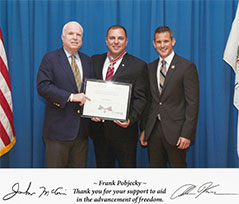 A group of three people standing together. The man in the middle is holding a Badge of Bravery award.