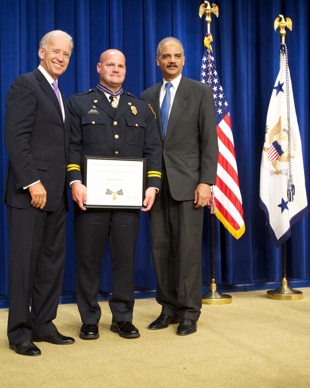 Three men standing in front of flags on a stage and facing the camera. The man in the middle is wearing an officer uniform and holding a Medal of Valor award plaque. 