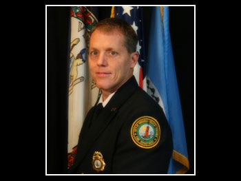 a man in a firefighter captain uniform standing in front of three flags and smiling