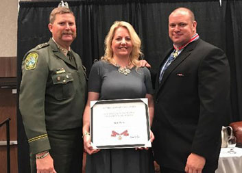 A group of three people standing together. A woman in the middle is holding a Badge of Bravery award.