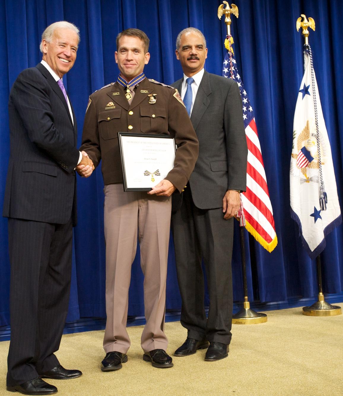 Three men standing in front of flags on a stage and facing the camera. The man in the middle is wearing an officer uniform and holding a Medal of Valor award plaque. 
