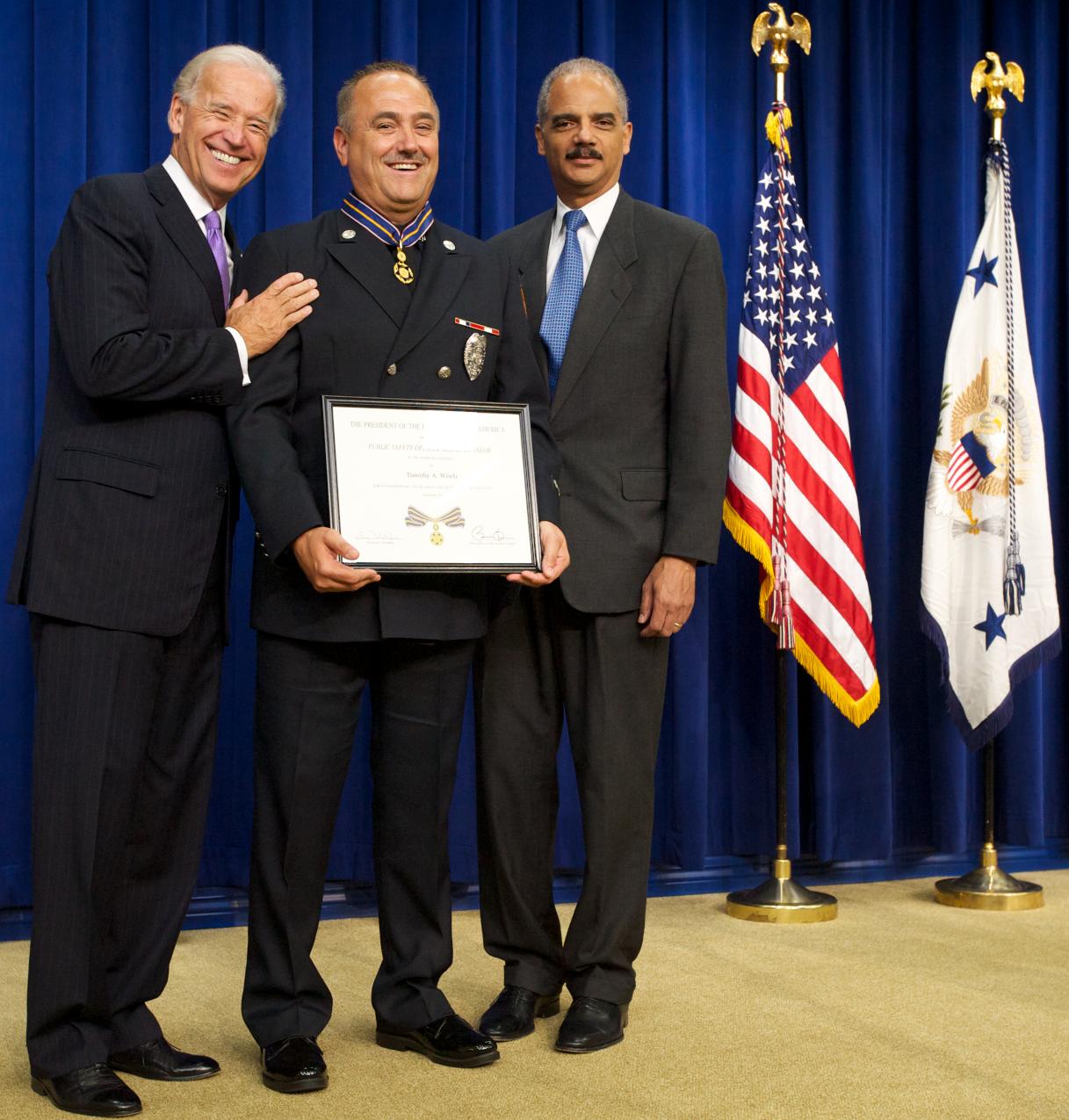 Three men standing in front of flags on a stage and smiling at the camera. The man in the middle is wearing an officer uniform and holding a Medal of Valor award plaque.
