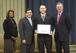 Four people in professional attire standing on a stage and smiling at the camera. One man is holding a Medal of Valor award plaque.