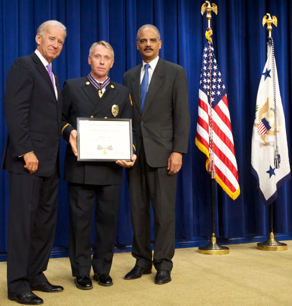 Three men standing in front of flags on a stage and facing the camera. The man in the middle is wearing an officer uniform and holding a Medal of Valor award plaque.