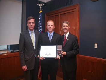A group of three people standing together. The man in the middle is holding a Badge of Bravery award.