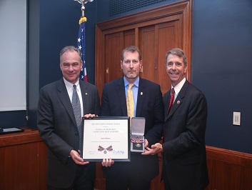 A group of three people standing together. The man in the middle is holding a Badge of Bravery award.