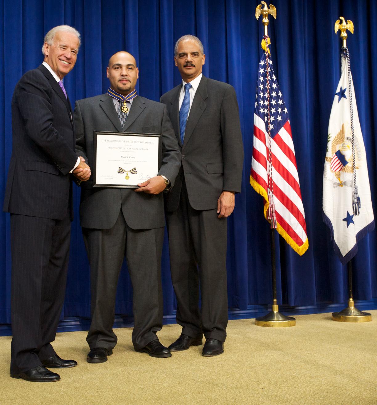 Three men wearing suits, standing in front of flags on a stage, and smiling at the camera. The man in the middle is holding a Medal of Valor award plaque.