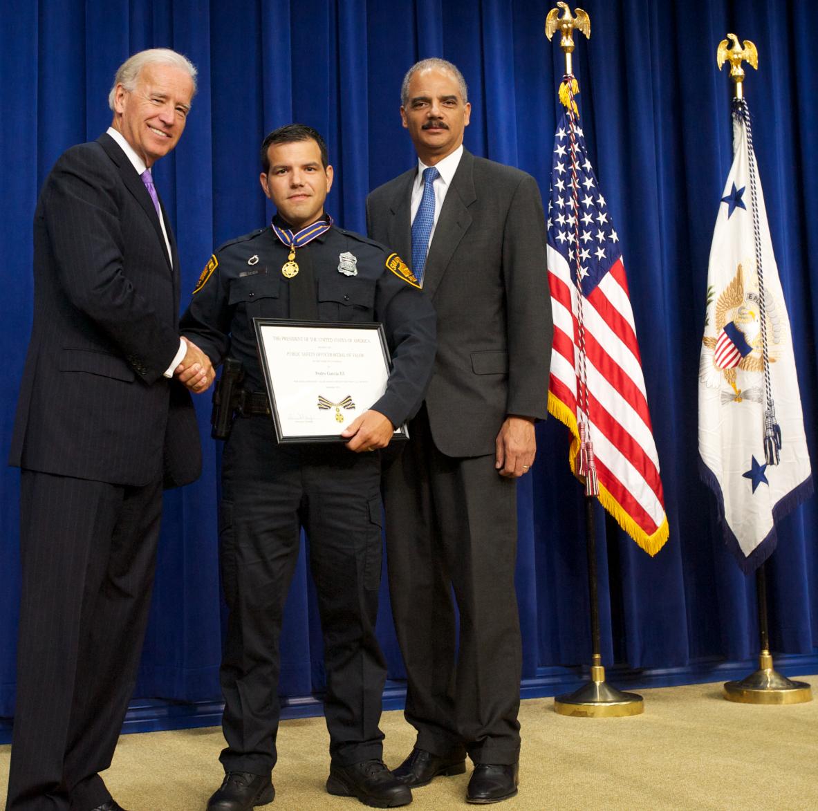 Three men standing in front of flags on a stage and facing the camera. The man in the middle is wearing an officer uniform and holding a Medal of Valor award plaque. 