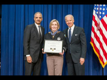 Two men and a woman standing on stage. The woman is in the middle and holding a Medal of Valor award plaque,