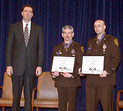Three men standing on a stage and facing the camera. Two of the men are wearing officer uniforms and holding a Medal of Valor award plaque.