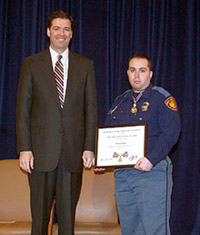 Two men standing on a stage and facing the camera. One man is wearing an officer uniform and holding a Medal of Valor award plaque.
