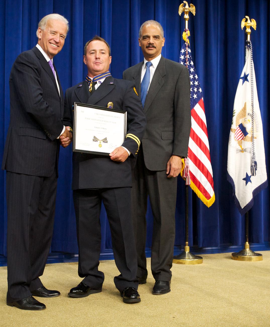 Three men standing in front of flags on a stage and facing the camera. The man in the middle is wearing an officer uniform and holding a Medal of Valor award plaque. 