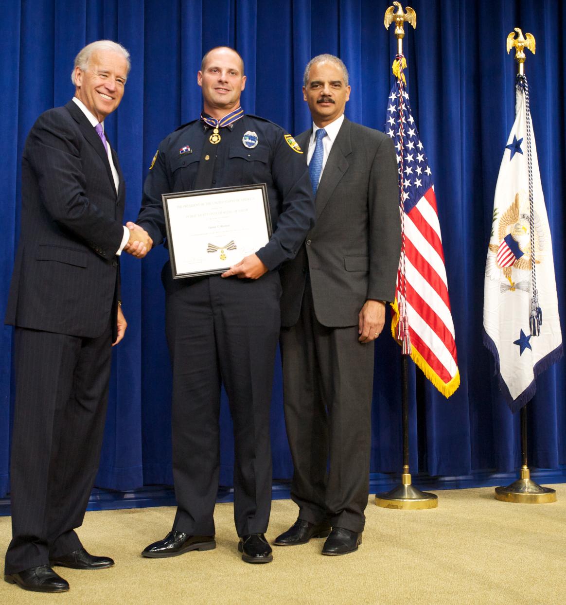 Three men standing in front of flags on a stage and facing the camera. The man in the middle is wearing an officer uniform and holding a Medal of Valor award plaque.