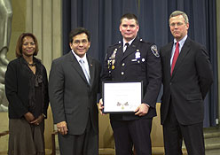 Four people standing on a stage, and smiling at the camera. One man is wearing an officer uniform and holding a Medal of Valor award plaque.