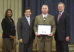 Four people wearing professional attire, standing on a stage, and smiling at the camera. One man is holding a Medal of Valor award plaque.