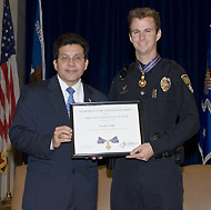 Two men standing on a stage and smiling at the camera. One man is in an officer uniform and the men are holding a Medal of Valor award plaque between them.