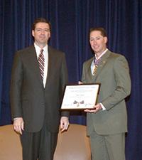 Two men wearing suits, standing on a stage, and smiling at the camera. One man is holding a Medal of Valor award plaque.