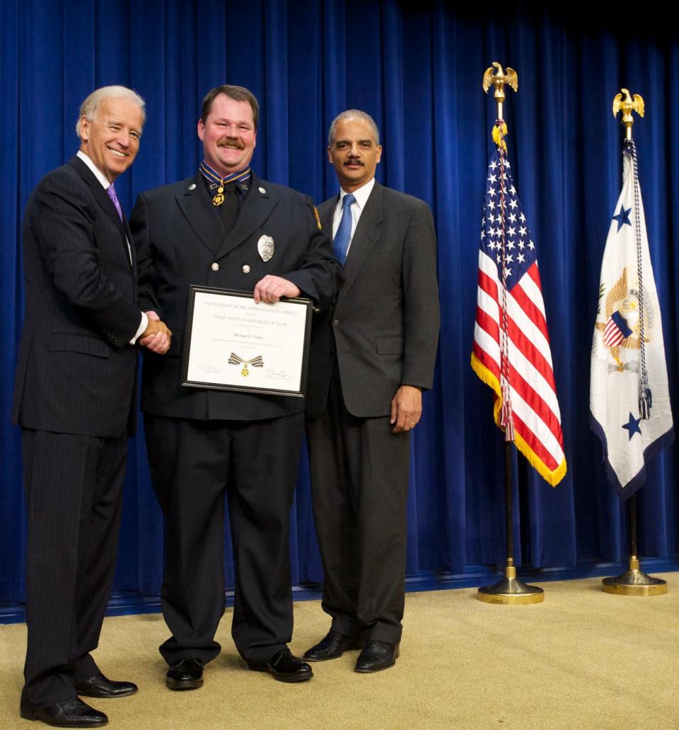 Three men standing in front of flags on a stage and smiling at the camera. The man in the middle is wearing an officer uniform and holding a Medal of Valor award plaque.