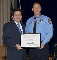 Two men standing on a stage and smiling at the camera. One man is in an officer uniform and is holding a Medal of Valor award plaque.