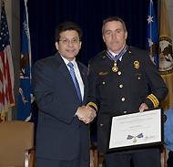 Two men standing on a stage, shaking hands, and smiling at the camera. One man is in an officer uniform and is holding a Medal of Valor award plaque.