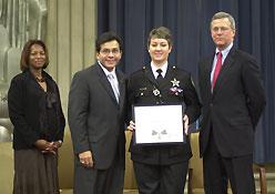 Four people standing on a stage and smiling at the camera. One woman is in an officer uniform and is holding a Medal of Valor award plaque.