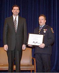 Two men standing on a stage and smiling at the camera. One man is wearing an officer uniform and holding a Medal of Valor award plaque.