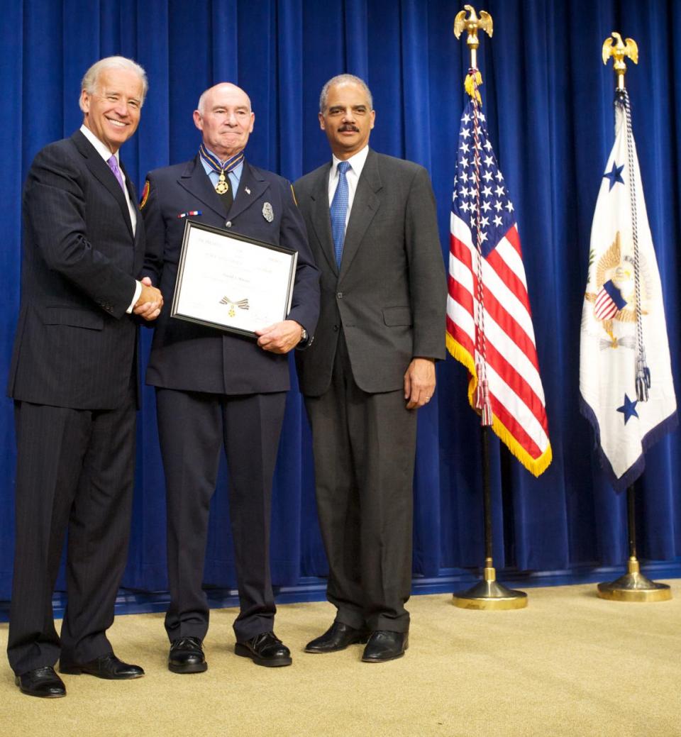 Three men standing in front of flags on a stage and smiling at the camera. The man in the middle is wearing an officer uniform and holding a Medal of Valor award plaque. 
