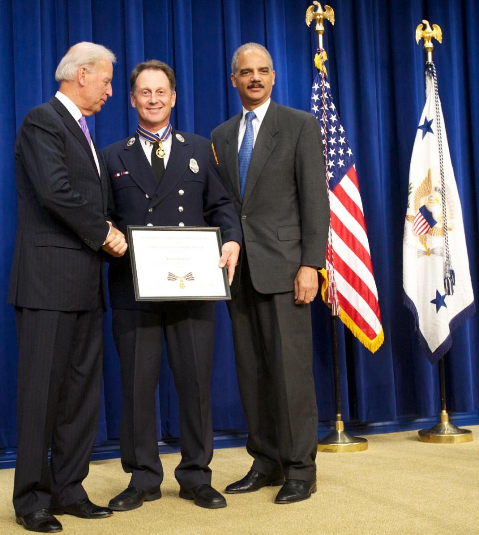 Three men standing together in front of flags on a stage. The man in the middle is wearing an officer uniform and holding a Medal of Valor award plaque. 