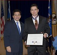Two men in suits standing on a stage and smiling at the camera. One man is holding a Medal of Valor award plaque.