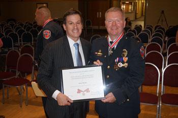 Two people standing together at the Badge of Bravery awards ceremony, holding an award between them, and smiling at the camera